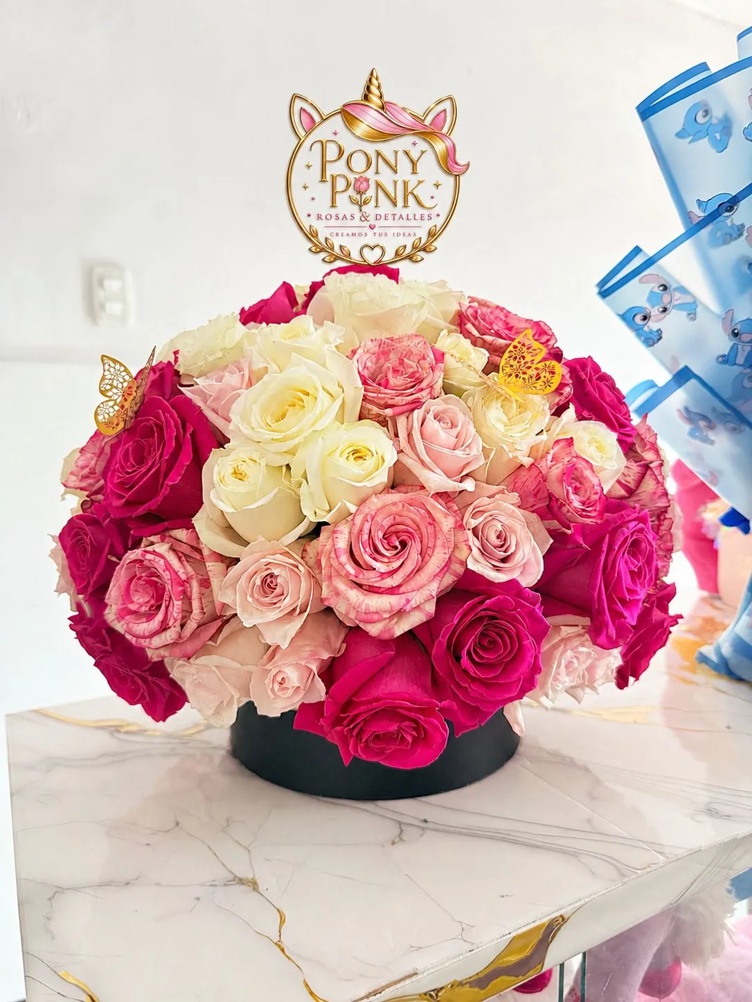 Arreglo circular de rosas fucsia, rosadas, crema y blancas montado en una base negra, con un cartel dorado de PonyPink y una mariposa.