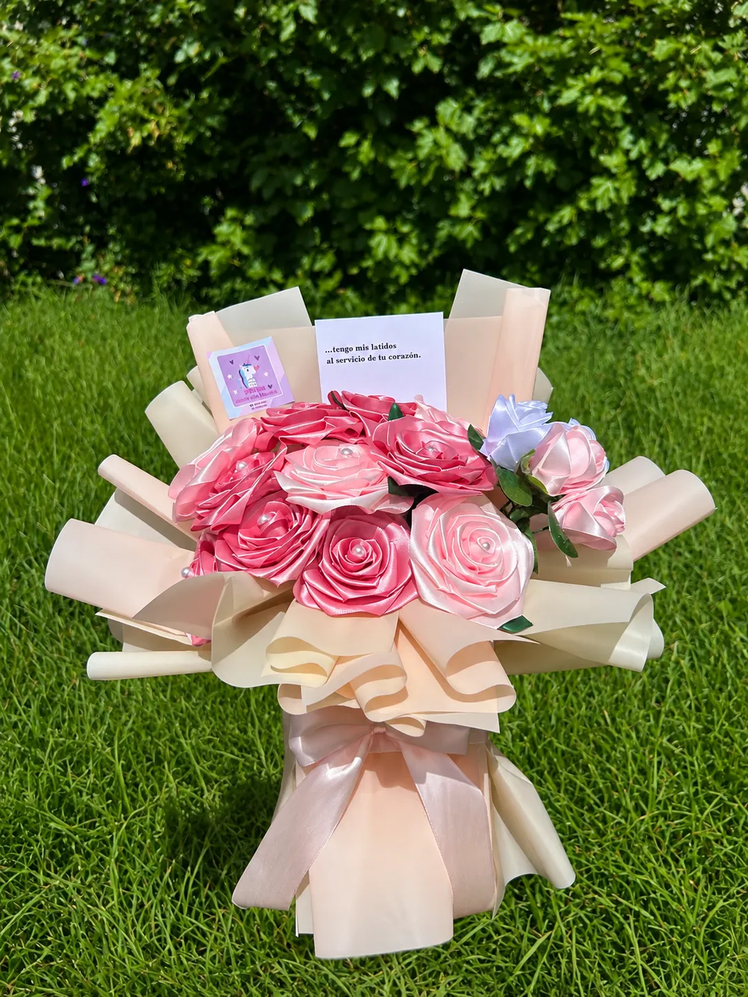 Ramo de rosas de cinta rosadas con flores azules y una tarjeta, en envoltura crema con lazo.