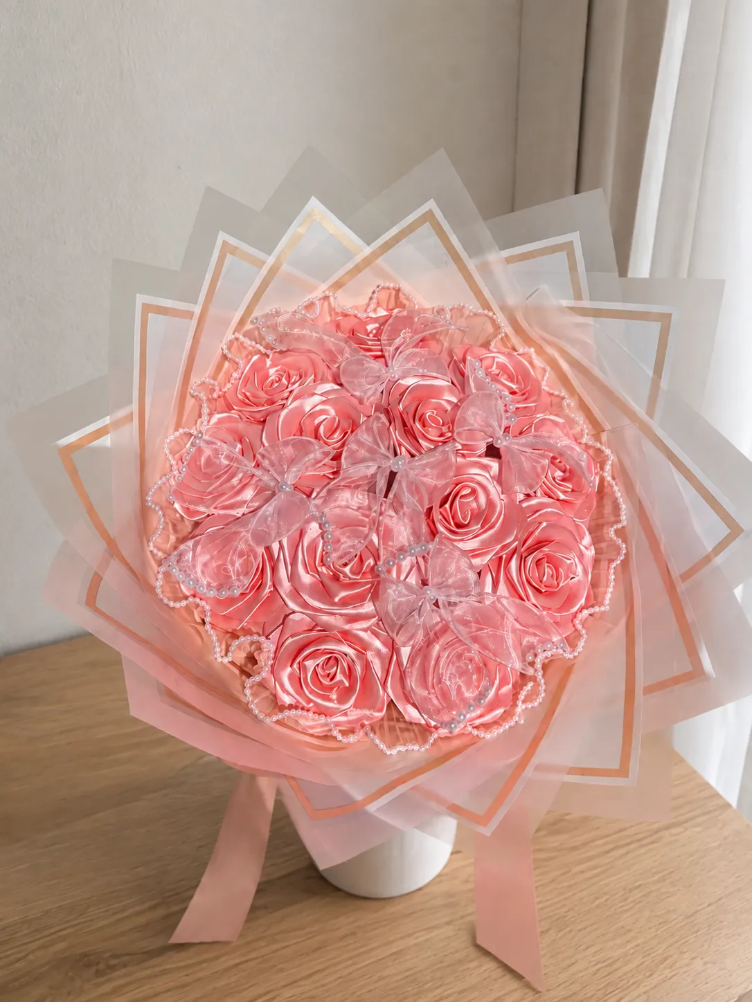 Ramo de rosas de cinta en tono durazno con envoltura translúcida y cintas, sobre una mesa de madera.