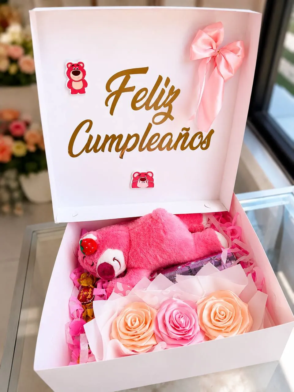 Caja rosa de cumpleaños abierta, con un peluche de oso, rosas de tela y golosinas.