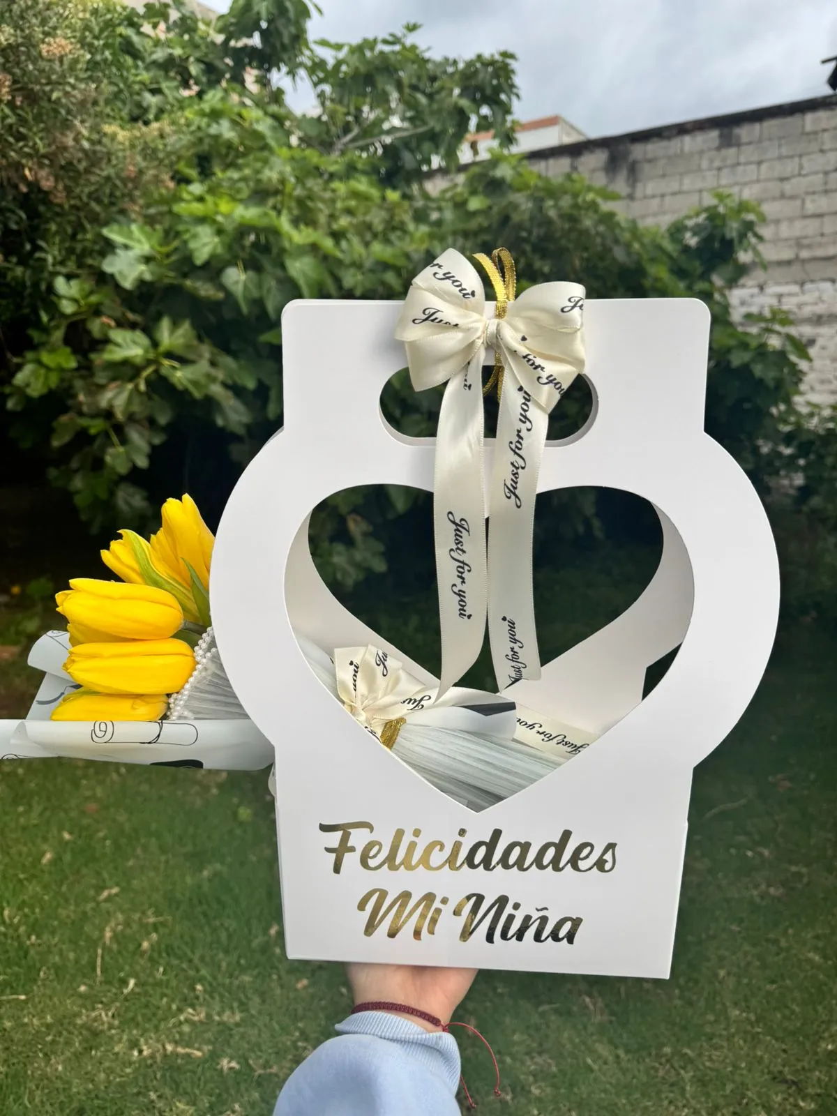 Caja blanca con una ventana recortada en forma de corazón, lazo de cinta crema y la frase «Felicidades mi niña» en dorado, junto a tulipanes amarillos.