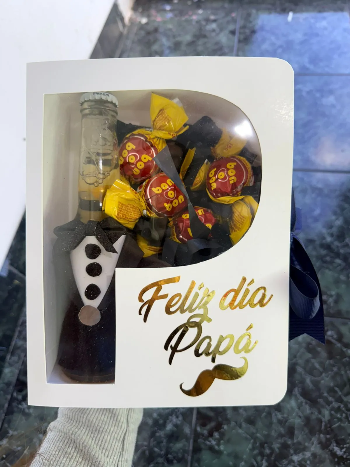 Caja blanca con forma de letra P y ventana transparente, con una botella pequeña vestida de esmoquin, bombones y la frase «Feliz día papá» en dorado.