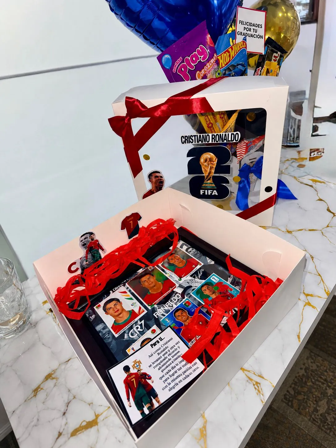 Caja abierta con cromos de fútbol y un adorno de futbolista sobre viruta roja, junto a otra caja con una lámina deportiva, globos azules y un cartel de graduación.