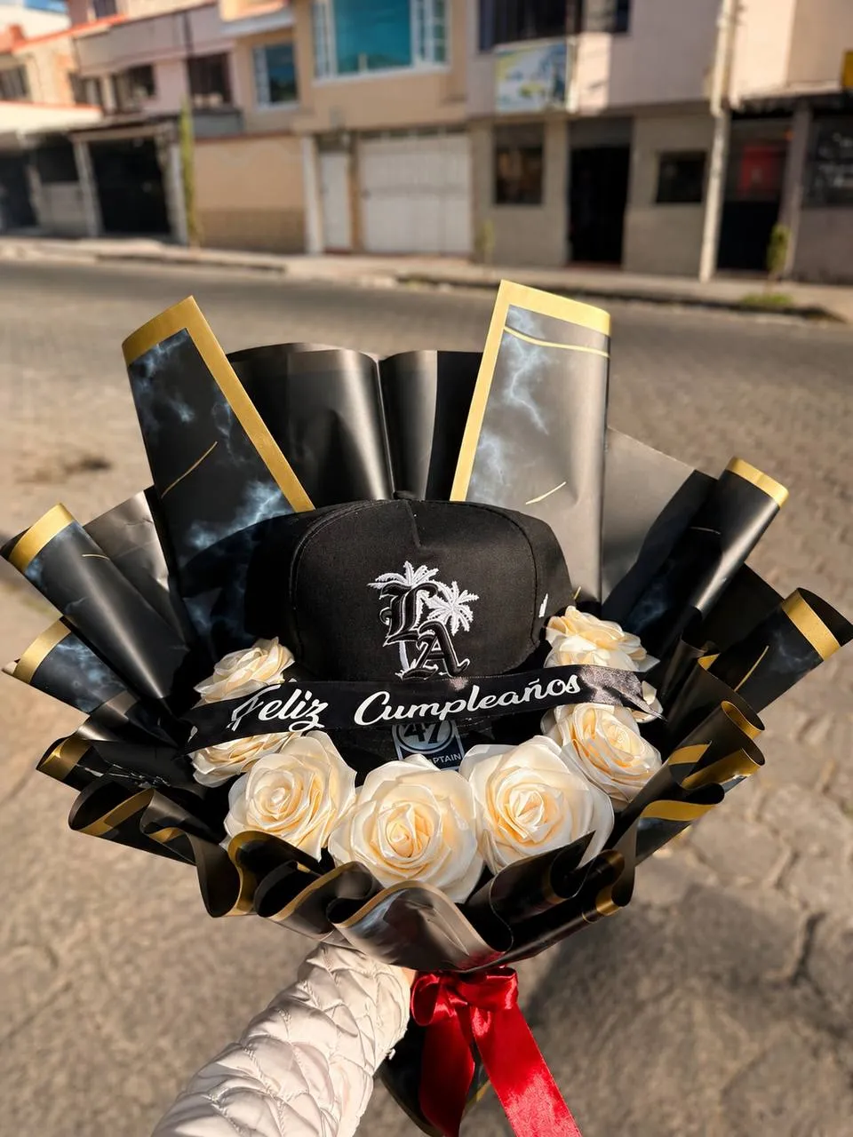 Ramo con una gorra negra de logotipo deportivo y rosas crema, con envoltura negra y dorada y banda de felicitación.