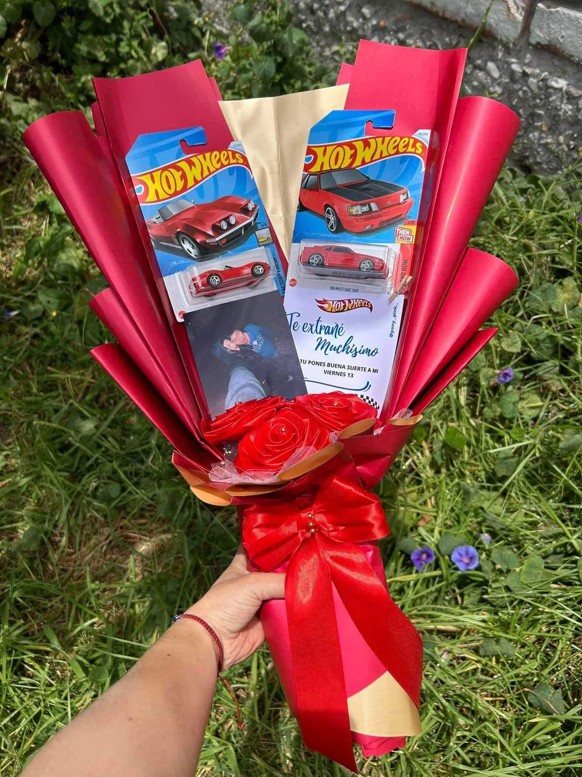 Ramo rojo con dos carros de juguete en su blíster, rosas de cinta rojas, una tarjeta con una dedicatoria y un lazo de raso.