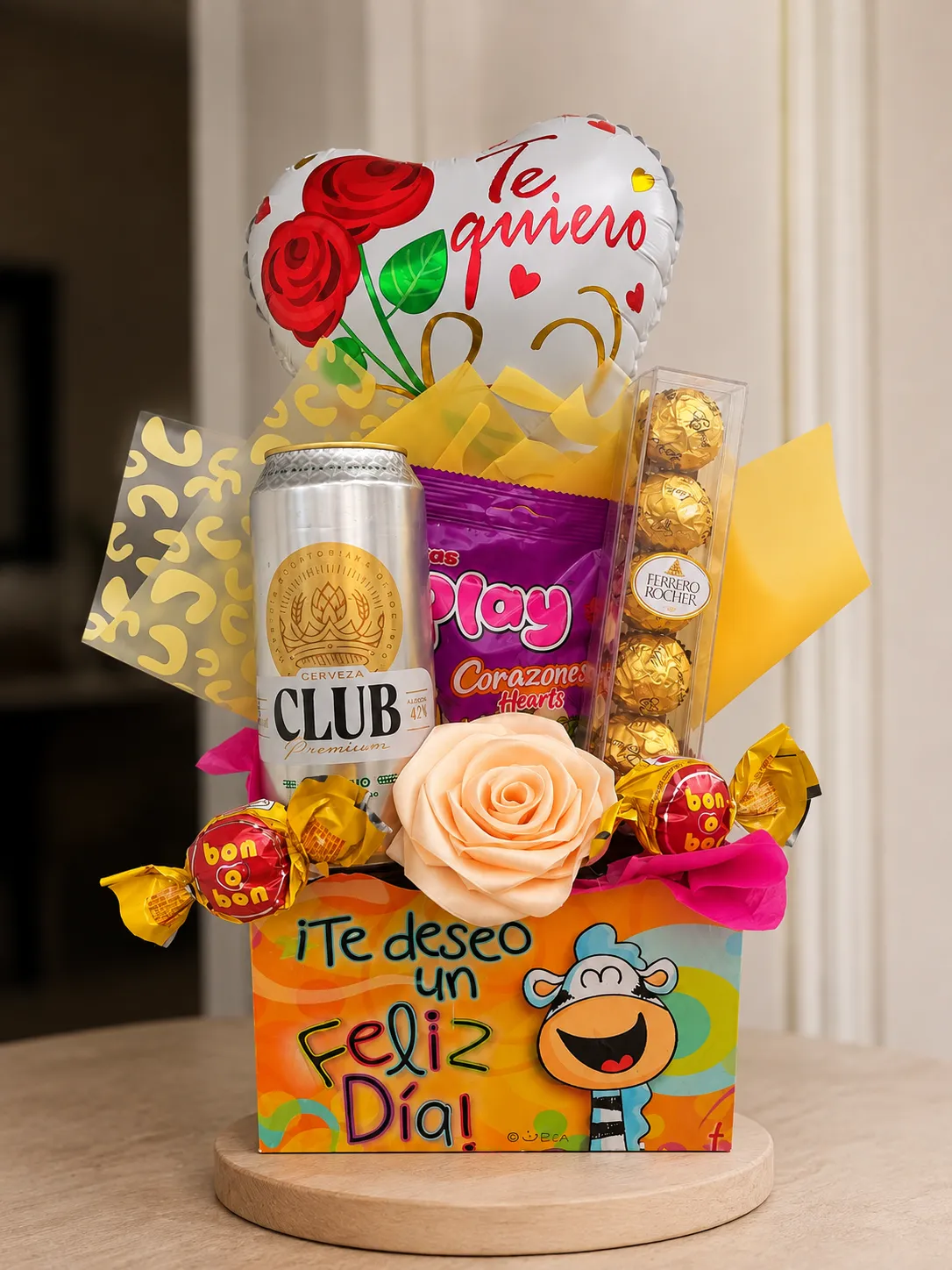 Caja colorida con un globo de corazón, una bebida, chocolates y una rosa de cinta.