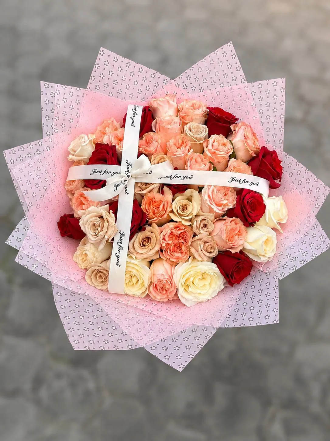 Ramo circular de rosas rosadas, durazno, crema y rojas con envoltura rosa estampada y cintas blancas.