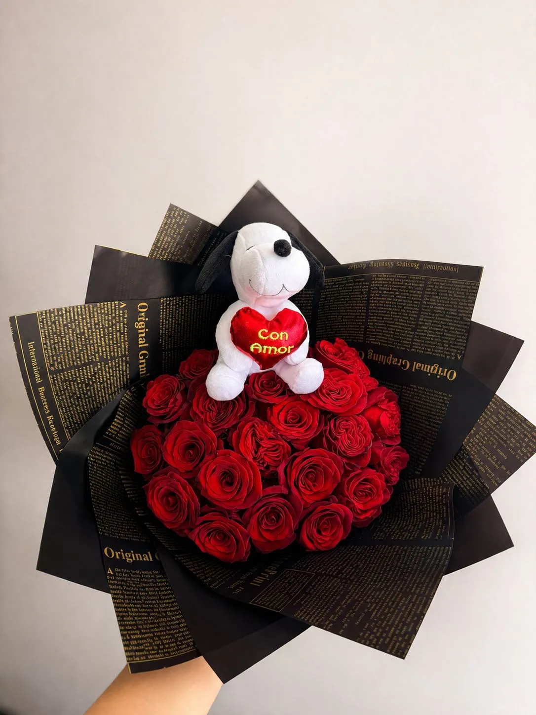 Ramo de rosas rojas con envoltura negra estampada de letras y un peluche blanco de perrito que sostiene un corazón rojo con la frase «Con amor».