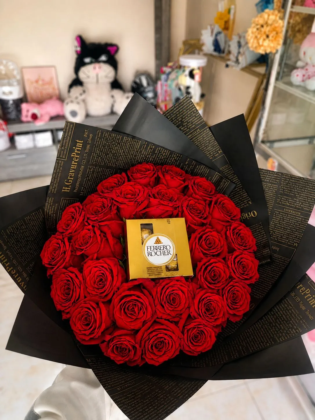 Ramo circular de rosas rojas con envoltura negra estampada de letras y una caja de bombones en el centro.