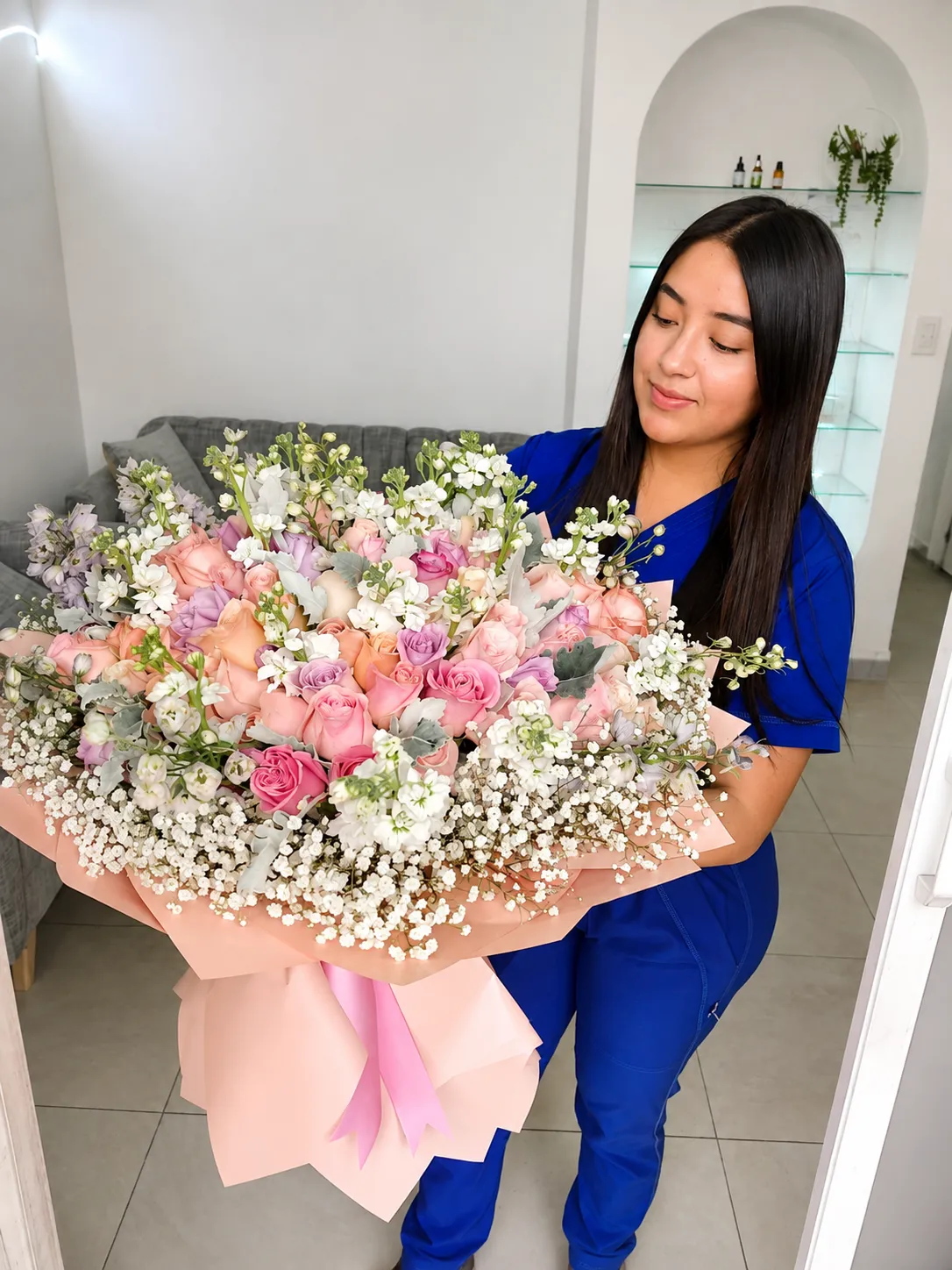 Una mujer vestida de azul mira un gran ramo de rosas rosadas, durazno y lilas con pequeñas flores blancas.