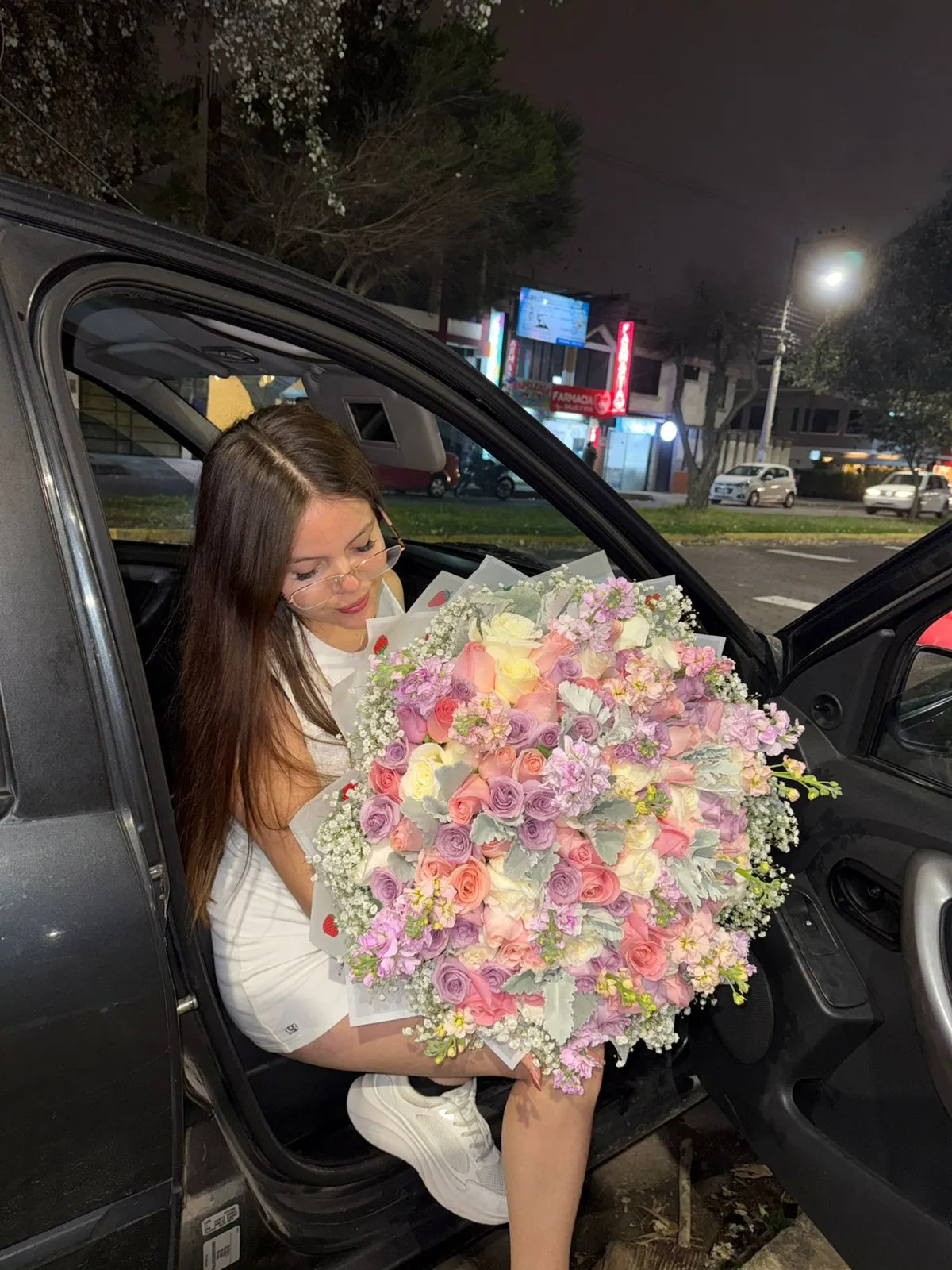 Una mujer sentada en un automóvil sostiene un ramo de rosas en tonos pastel y flores lilas durante la noche.
