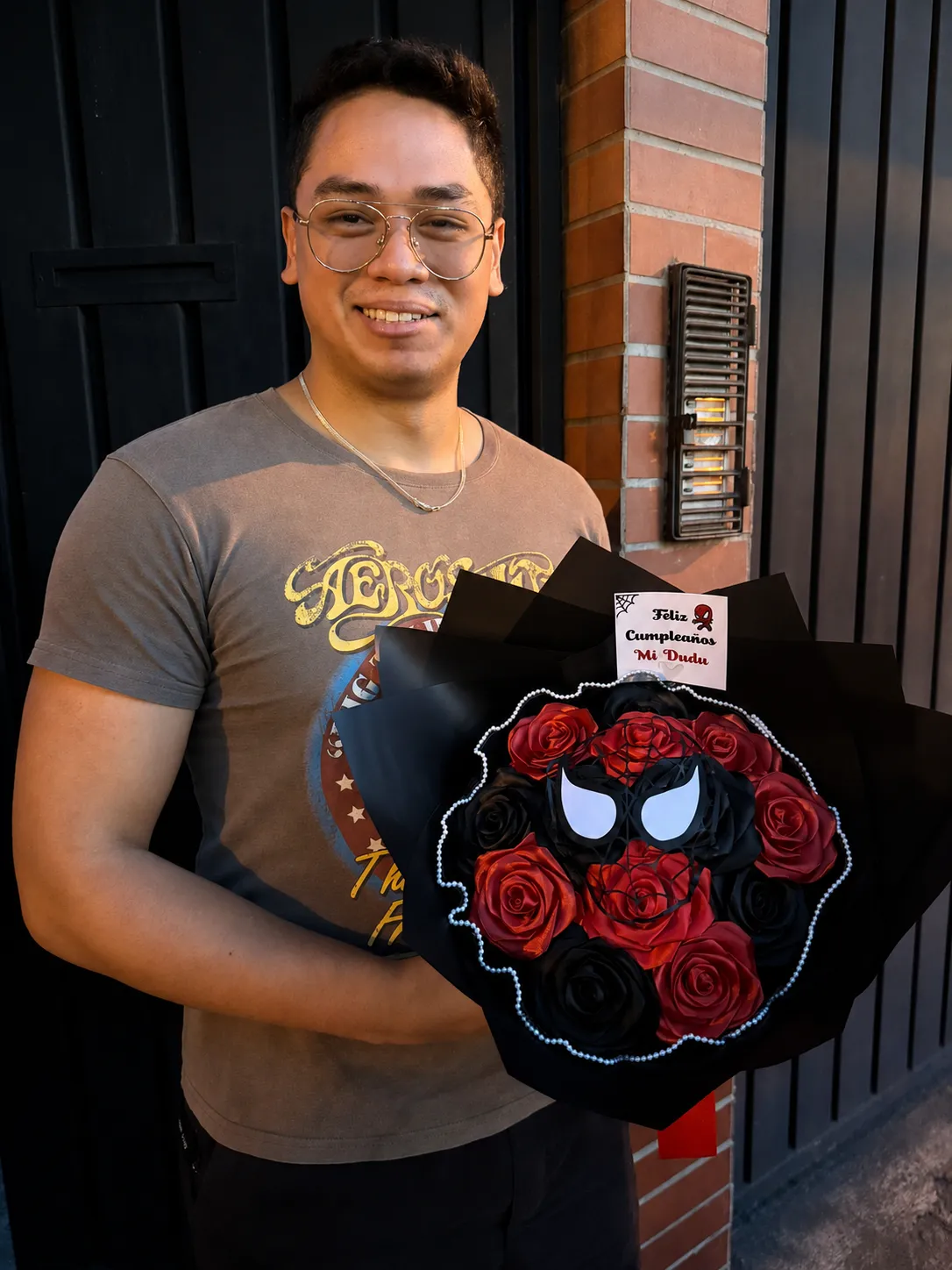 Un hombre sonríe con un ramo de rosas rojas y negras, un adorno de superhéroe y una tarjeta de cumpleaños.