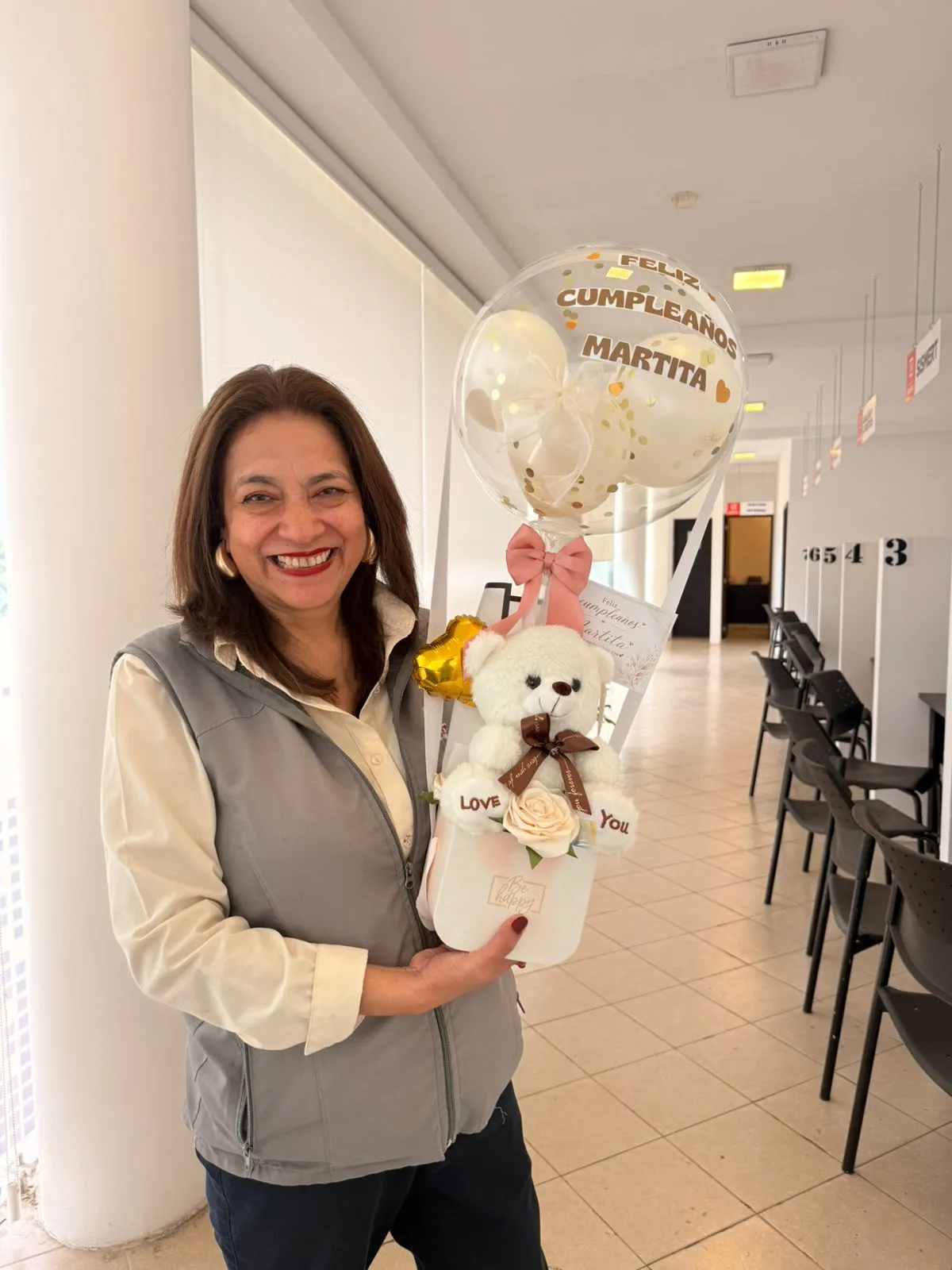 Una mujer sonríe con un arreglo de osito blanco, rosas y un globo transparente de cumpleaños.