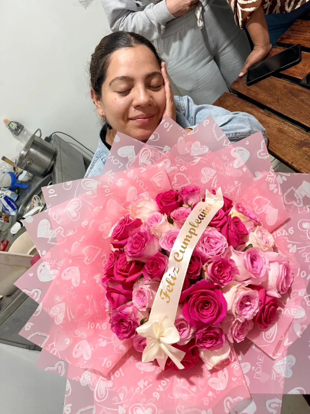 Una mujer sonríe con los ojos cerrados junto a un ramo de rosas rosadas y fucsias con una cinta de cumpleaños.