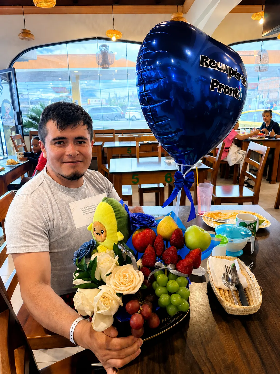 Un hombre sostiene un arreglo con rosas blancas y azules, frutas, un peluche de aguacate y un globo azul.
