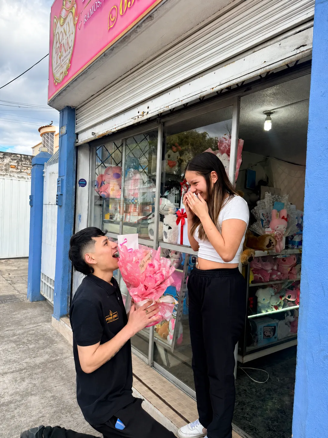 Un joven arrodillado ofrece un ramo de rosas rosadas a una joven que sonríe frente al local de PonyPink.