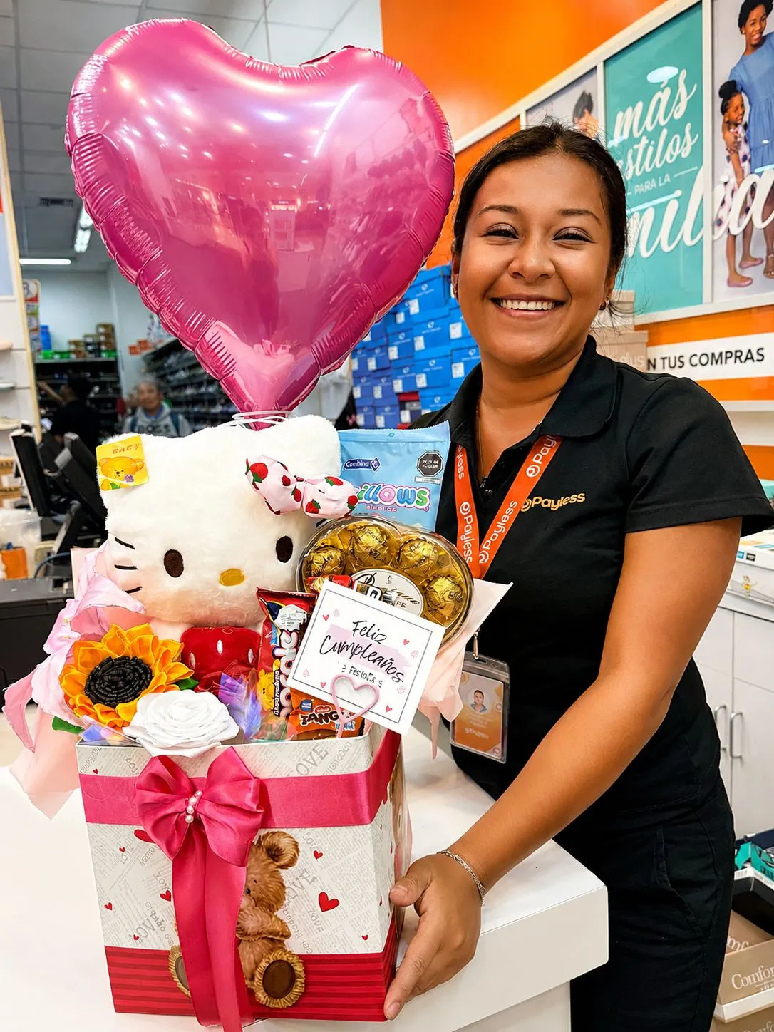 Una mujer sonríe sosteniendo una caja con un peluche blanco, chocolates, flores y un globo rosa de corazón.